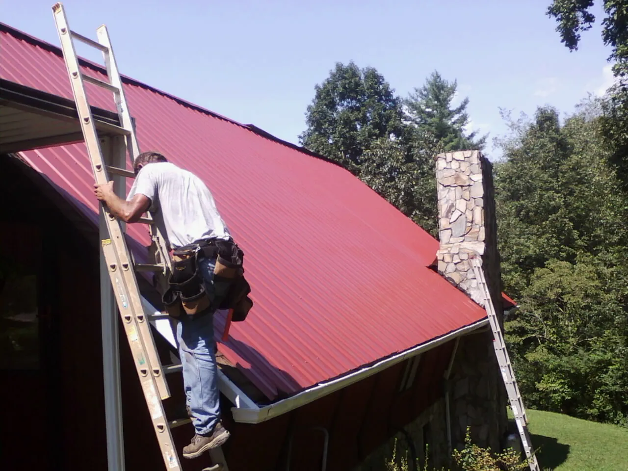 Roofer on a ladder beside a red metal roof and stone chimney — Soffit & Fascia Repair by Roofing Brothers in Huntington Beach